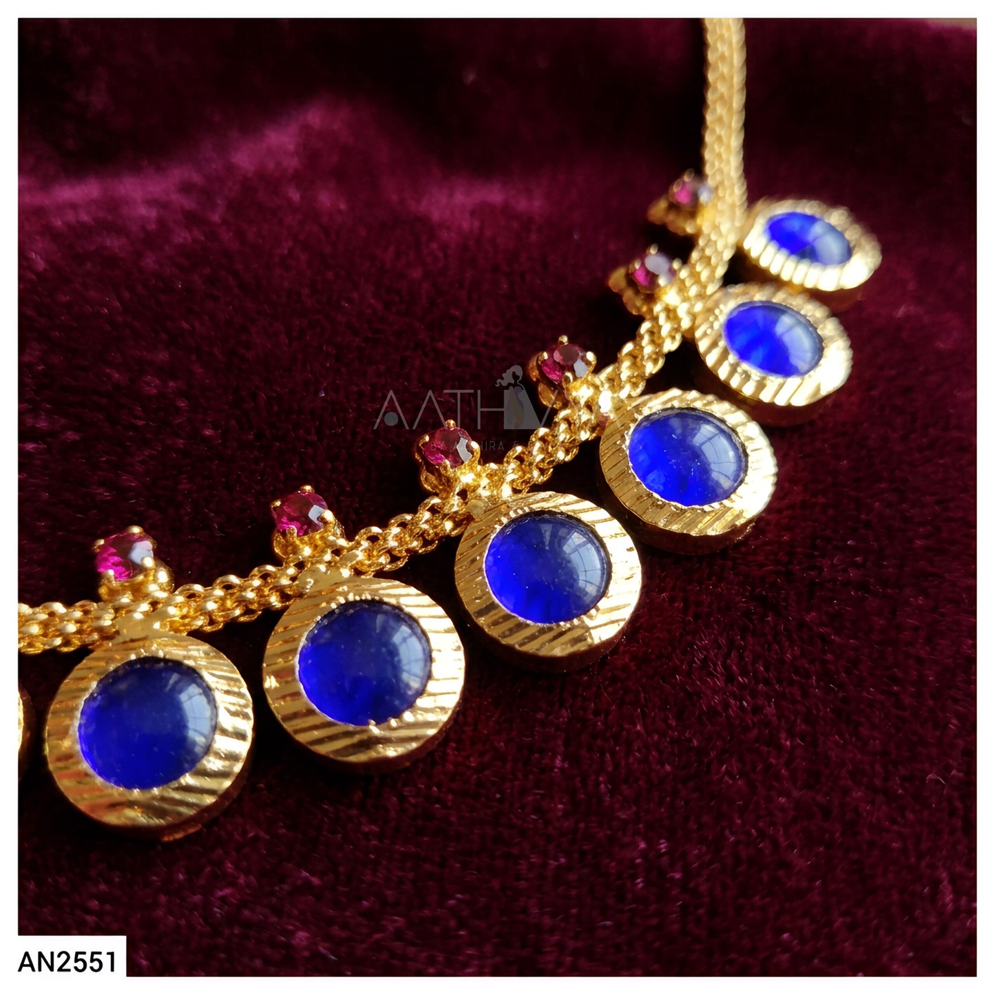 Gold chain necklace with five oval blue stones and small pink accent stones, displayed on a dark burgundy velvet background.