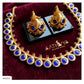 Gold jewelry set with a necklace and matching earrings, featuring large blue circular stones and small pink accent stones, displayed on a dark red velvet surface with the brand name AATHIVA visible on the box.