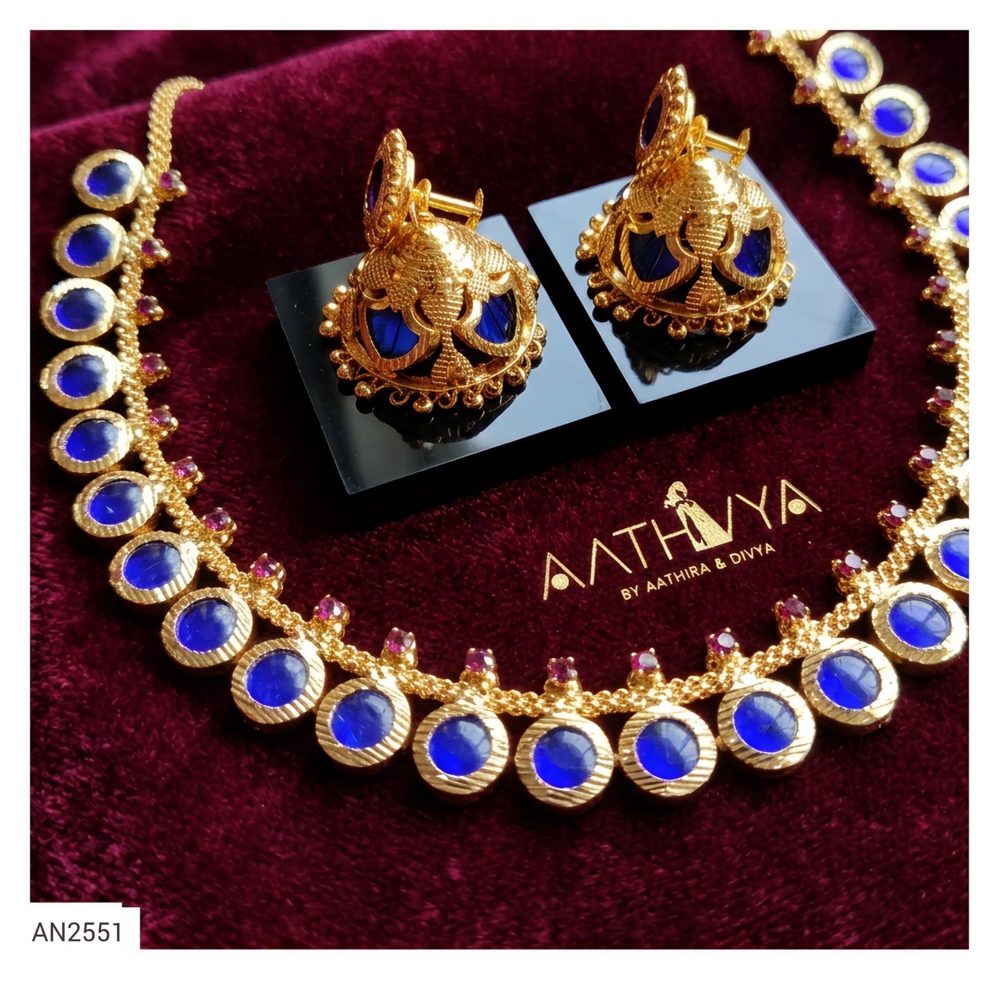 Gold jewelry set with a necklace and matching earrings, featuring large blue circular stones and small pink accent stones, displayed on a dark red velvet surface with the brand name AATHIVA visible on the box.