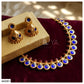 Gold-toned traditional Indian jewelry set including a necklace and matching earrings with deep blue oval stones and small red accent stones, displayed on a beige box and dark burgundy velvet surface.