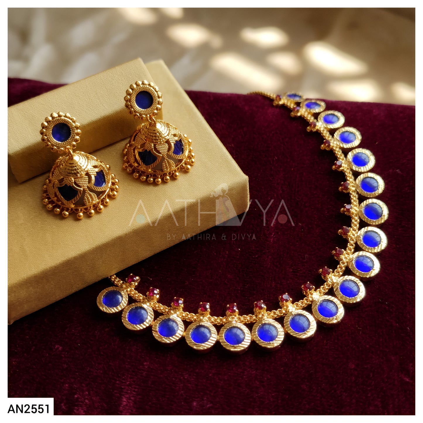 Gold-toned traditional Indian jewelry set including a necklace and matching earrings with deep blue oval stones and small red accent stones, displayed on a beige box and dark burgundy velvet surface.