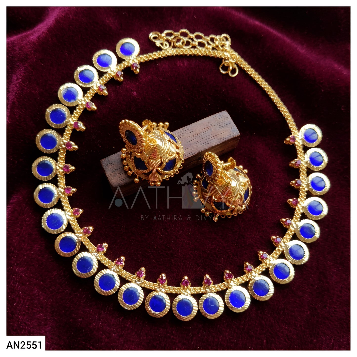 Gold-plated jewelry set featuring a necklace and matching earrings with round blue stones and small pink accent stones, displayed on a dark red velvet background.