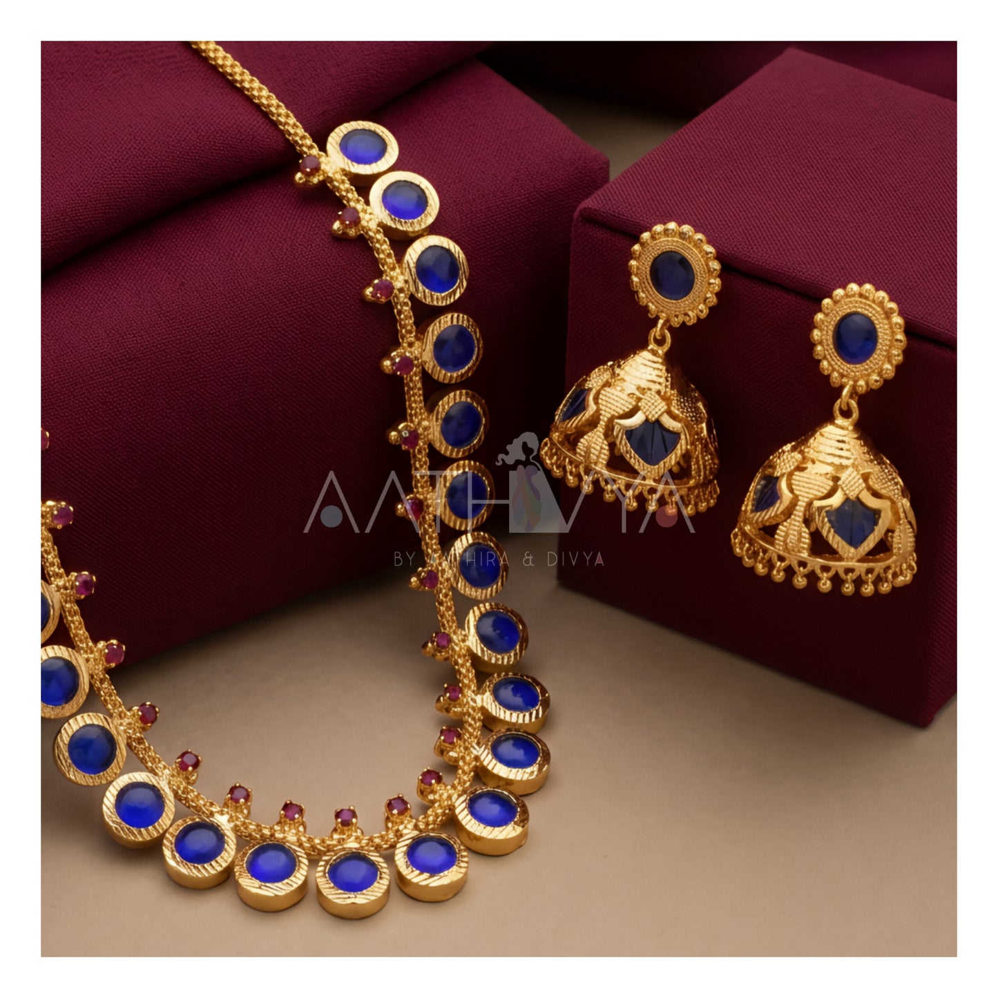 Gold jewelry set featuring a necklace and matching earrings with deep blue oval stones and small red accent stones, displayed on dark burgundy boxes against a beige background.