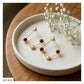 A pair of gold geometric dangle earrings with white pearl-like beads and red faceted gemstone accents, displayed on a white ceramic plate with baby's breath flowers on a wooden table.