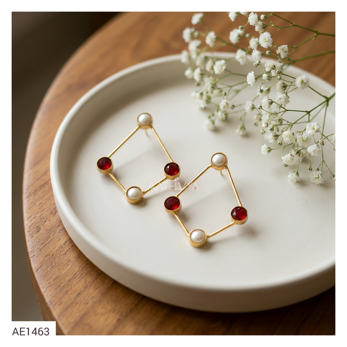 A pair of gold geometric dangle earrings with white pearl-like beads and red faceted gemstone accents, displayed on a white ceramic plate with baby's breath flowers on a wooden table.