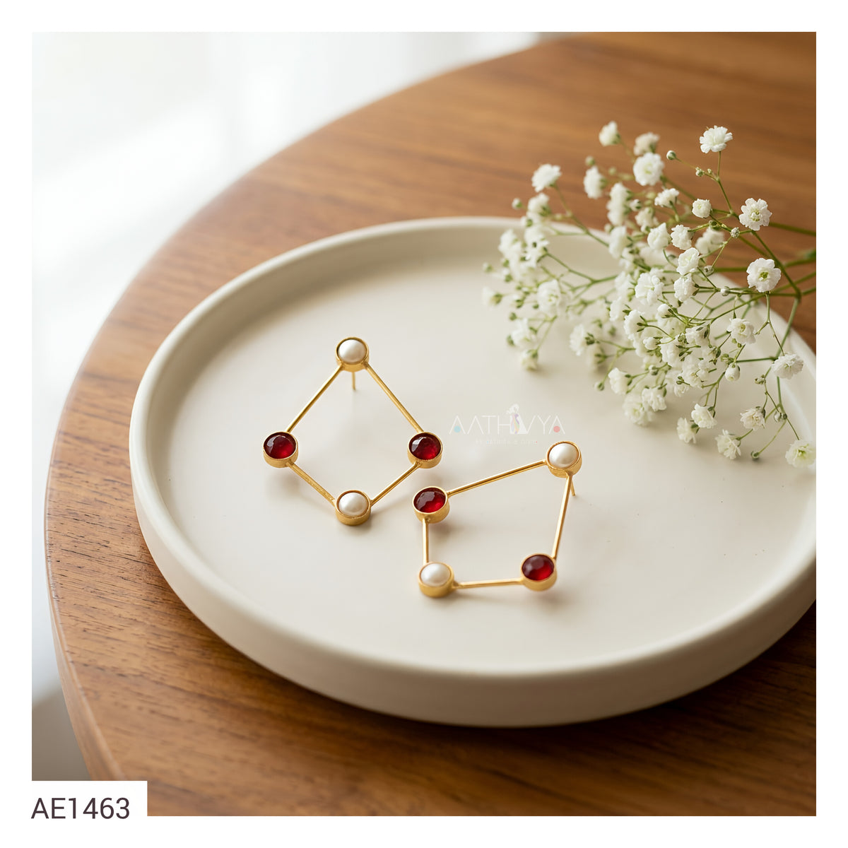 A pair of gold geometric stud earrings with red gemstone accents and pearl-like beads, displayed on a round cream dish on a wooden table with small white flowers.