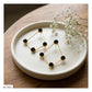 Gold geometric stud earrings with black textured circular centers, displayed on a round white ceramic plate with baby's breath flowers on a wooden table.