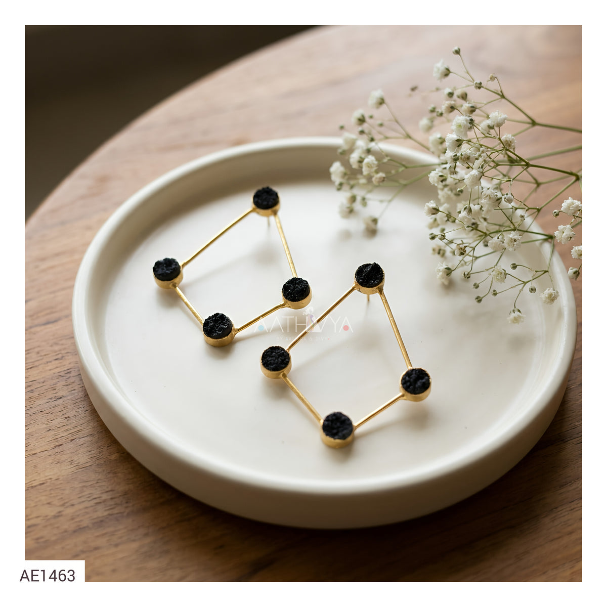 Gold geometric stud earrings with black textured circular centers, displayed on a round white ceramic plate with baby's breath flowers on a wooden table.