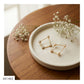 Gold geometric stud earrings with small pearl-like beads, displayed on a round white ceramic dish on a wooden table with baby's breath flowers.