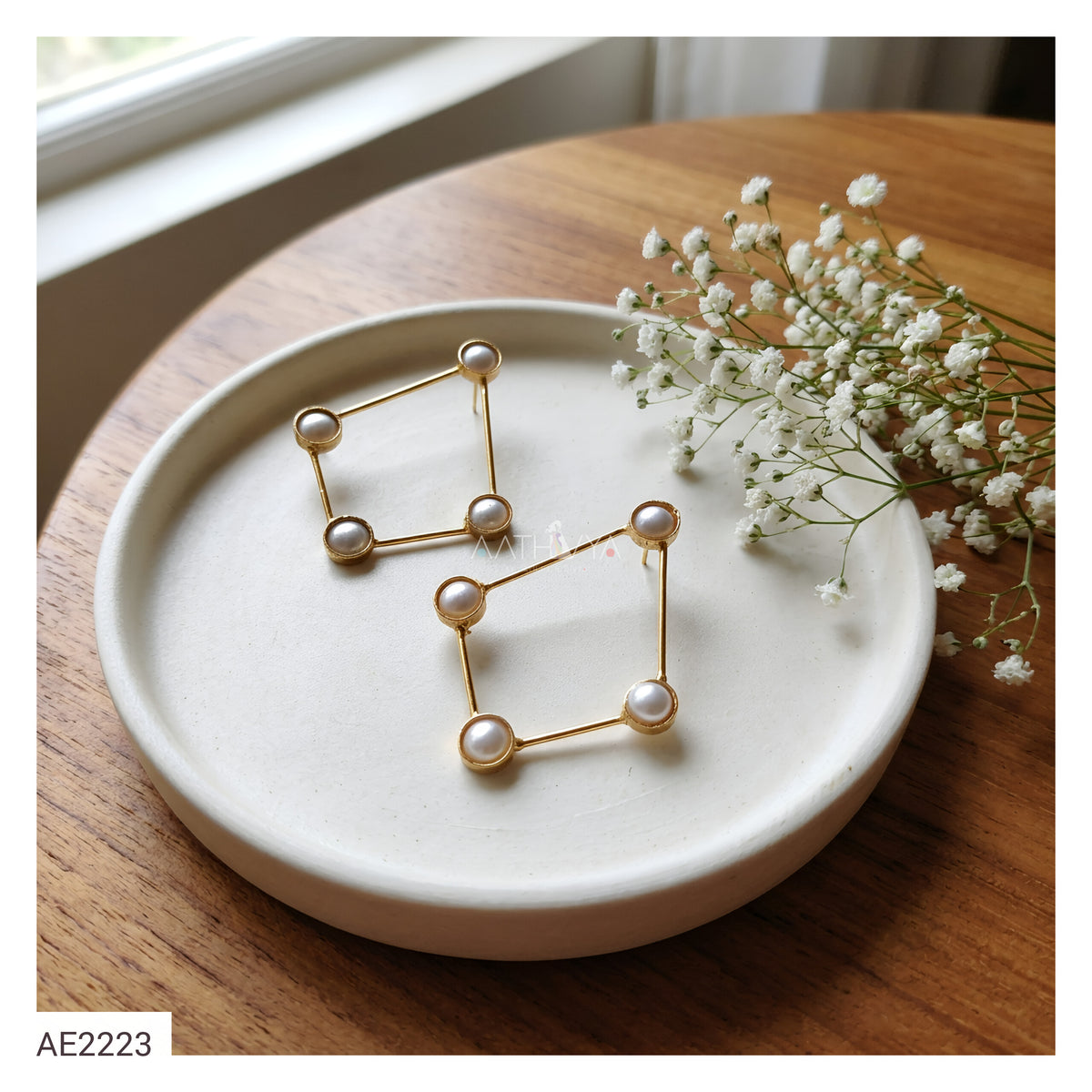 Two gold geometric stud earrings with white pearl-like beads, displayed on a round white ceramic dish on a wooden table with baby's breath flowers.