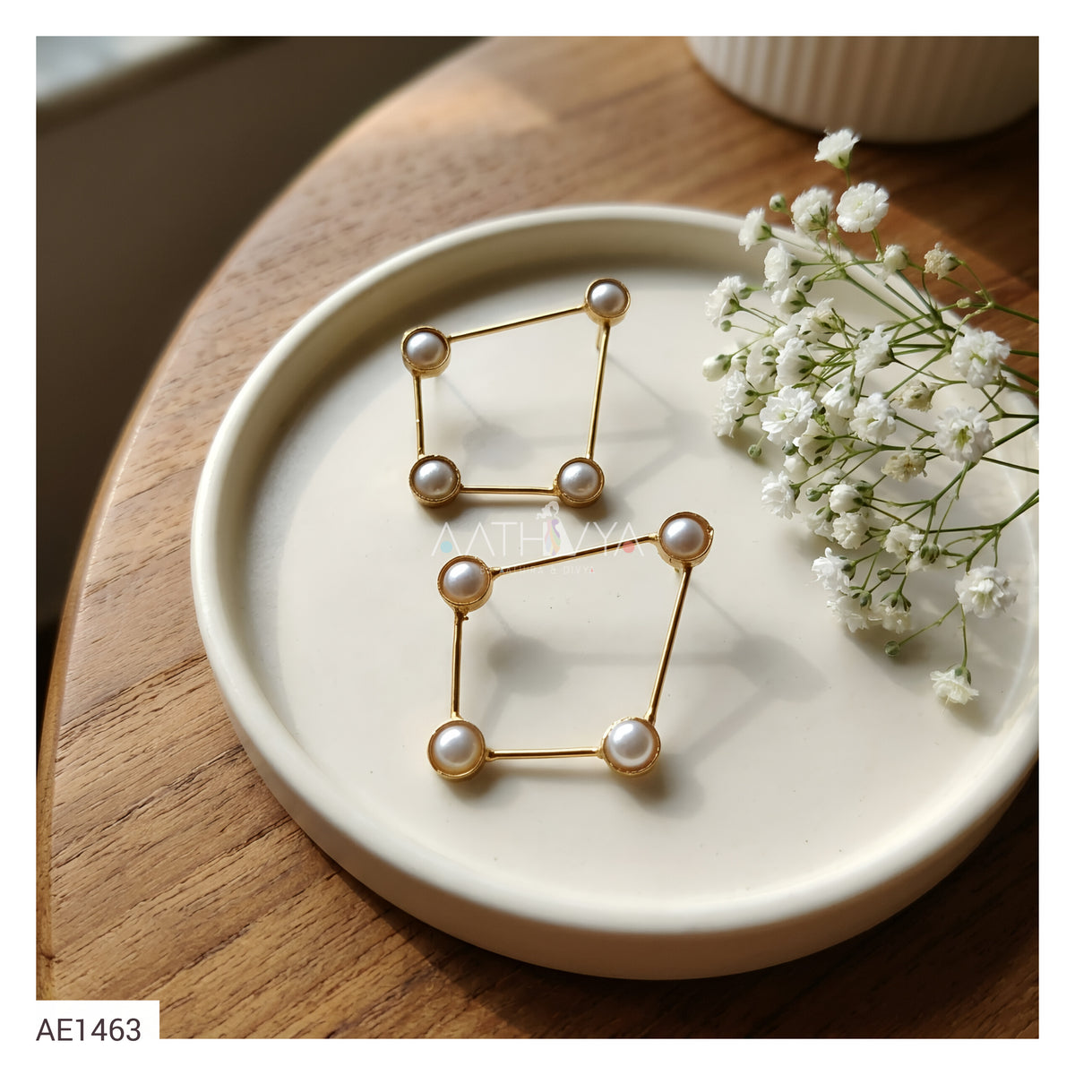 A pair of gold geometric stud earrings with white faux pearls at the corners, displayed on a round white ceramic plate with baby's breath flowers on a wooden table.