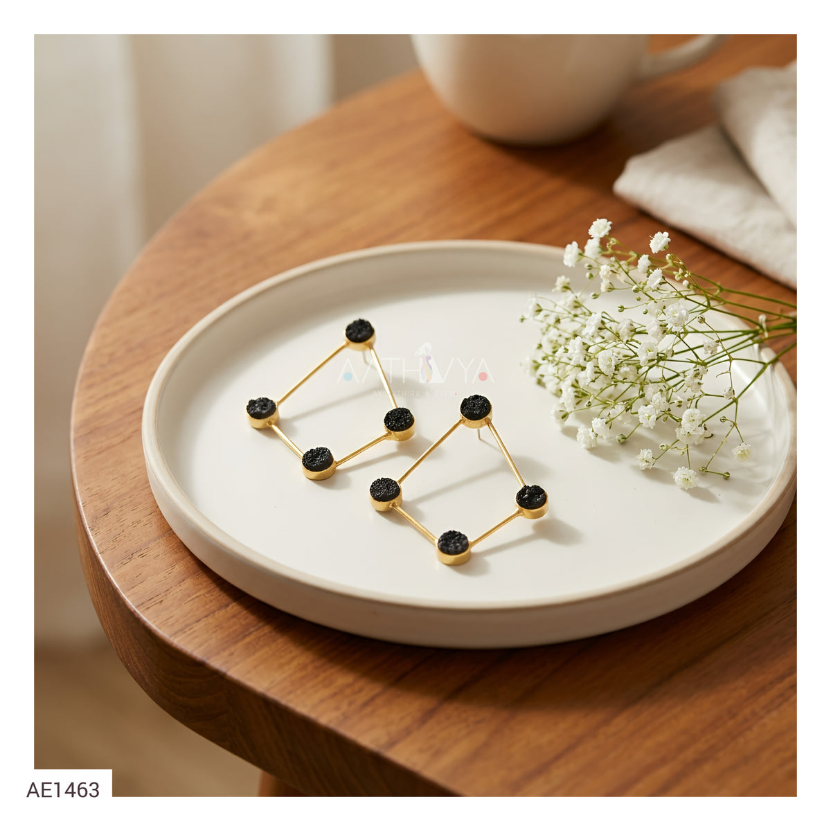 A pair of gold geometric triangle earrings with black circular studs, displayed on a round white ceramic plate on a wooden table next to baby's breath flowers.