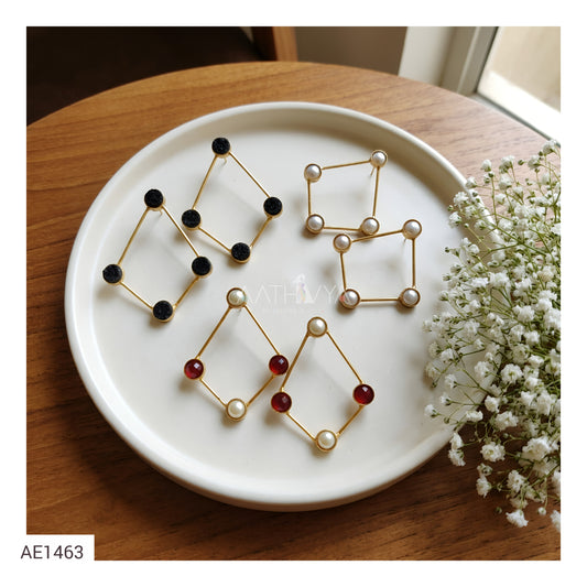 Three pairs of geometric gold-tone earrings displayed on a white ceramic plate, with black, pearl, and red bead accents, next to baby's breath flowers on a wooden table.