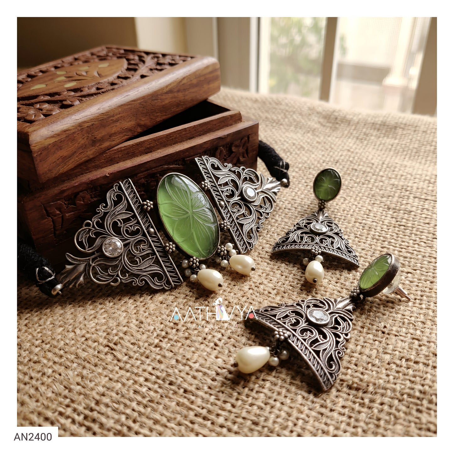 An ornate jewelry set displayed on a burlap surface next to a carved wooden box. The set includes a large triangular pendant with a green oval stone and silver filigree, matching drop earrings, and a black cord with a small white pearl bead.