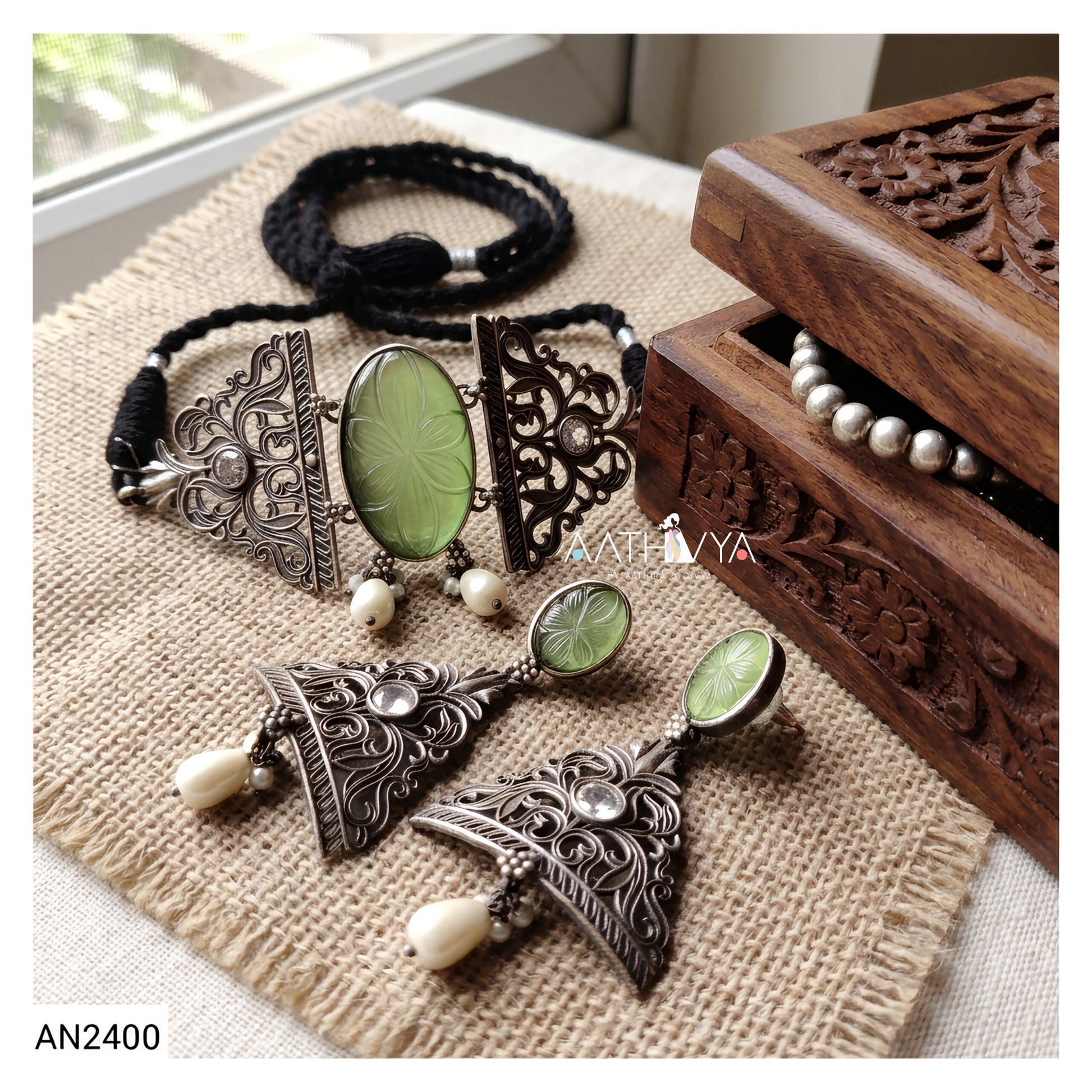 A jewelry set displayed on a beige burlap cloth next to a carved wooden box. The set includes a black braided bracelet, a silver-toned filigree necklace with oval green stone pendants and small white beads, matching silver-toned earrings, and a partial pearl necklace inside the wooden box.