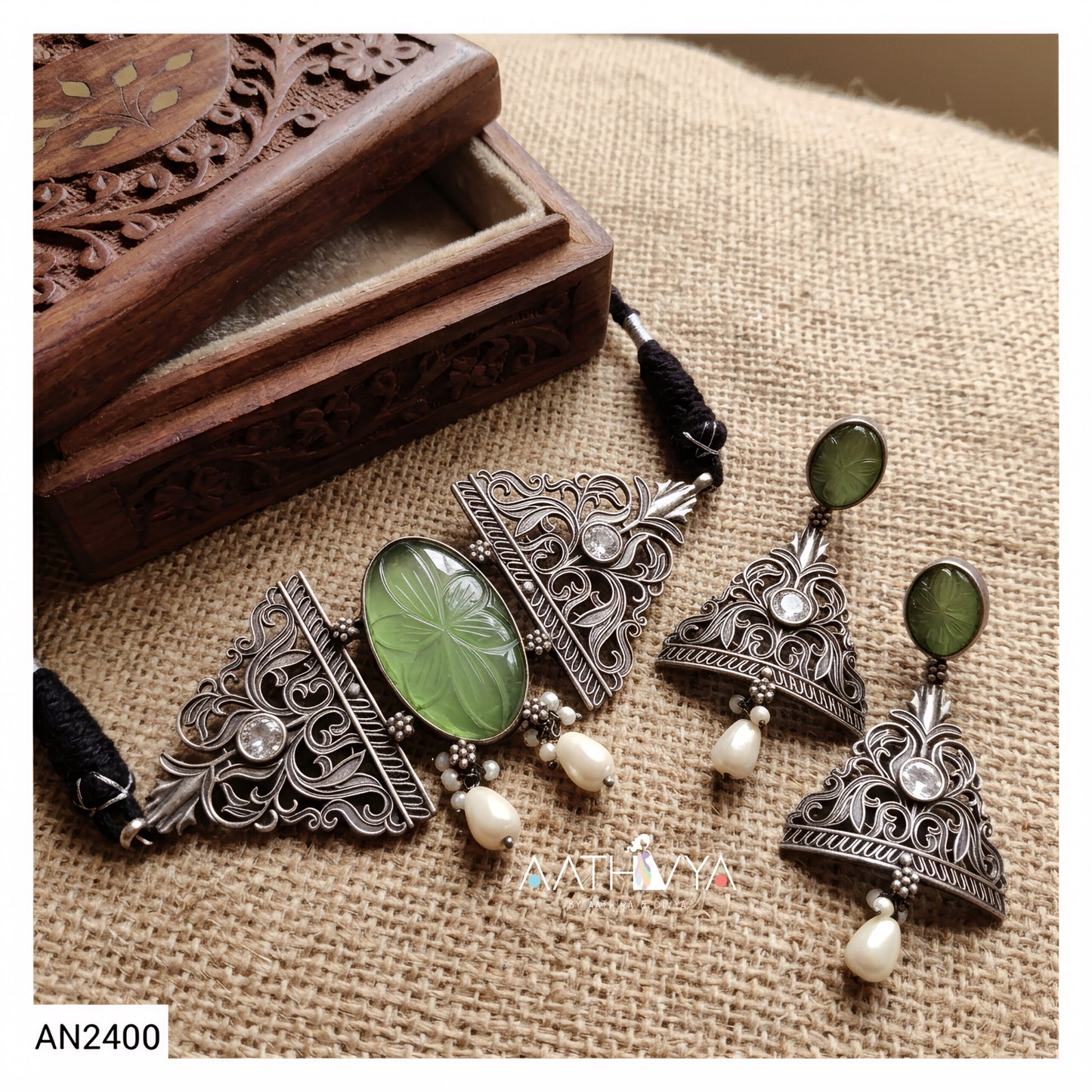 An antique-style silver-toned jewelry set featuring a necklace and matching earrings with green stone and pearl details, displayed on a burlap fabric next to a carved wooden box.