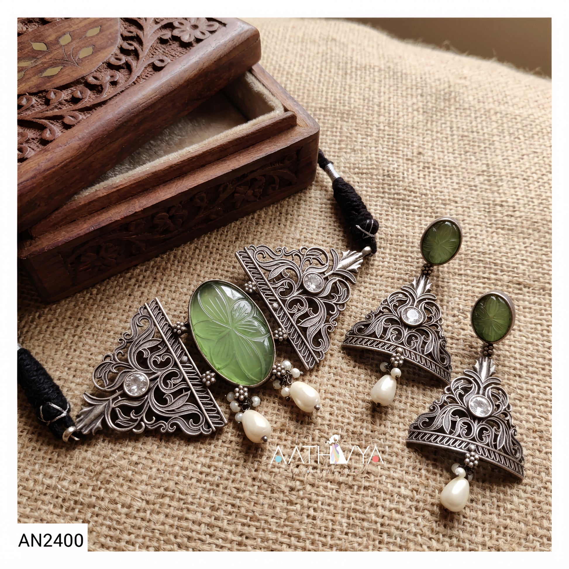An antique-style silver-toned jewelry set featuring a necklace and matching earrings with green stone and pearl details, displayed on a burlap fabric next to a carved wooden box.