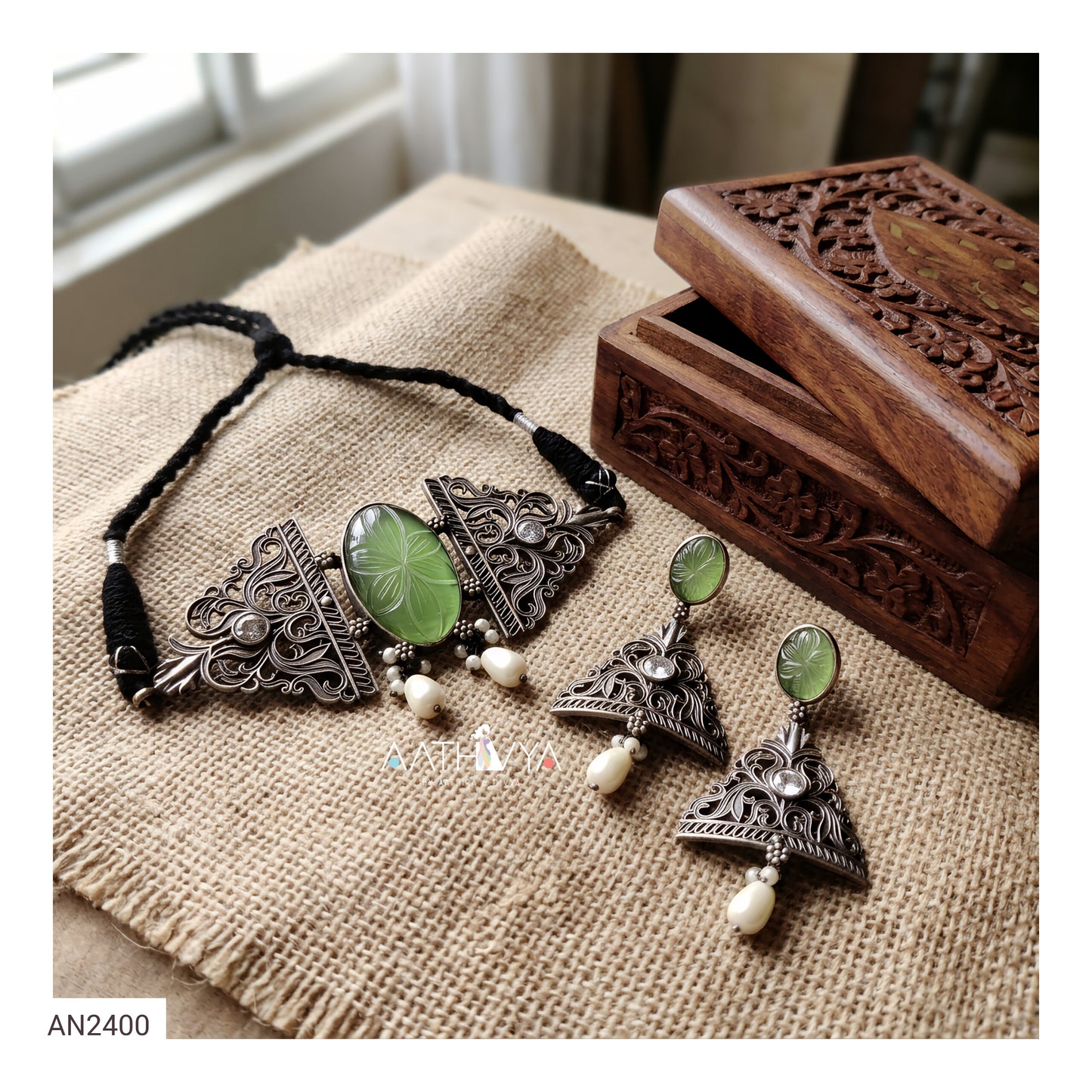 A jewelry set displayed on a burlap fabric surface next to a carved wooden box. The set includes a necklace with a black cord and three silver-toned filigree triangles with oval green stone centers, plus matching drop earrings with silver-toned filigree, green oval stones, and white bead drops.
