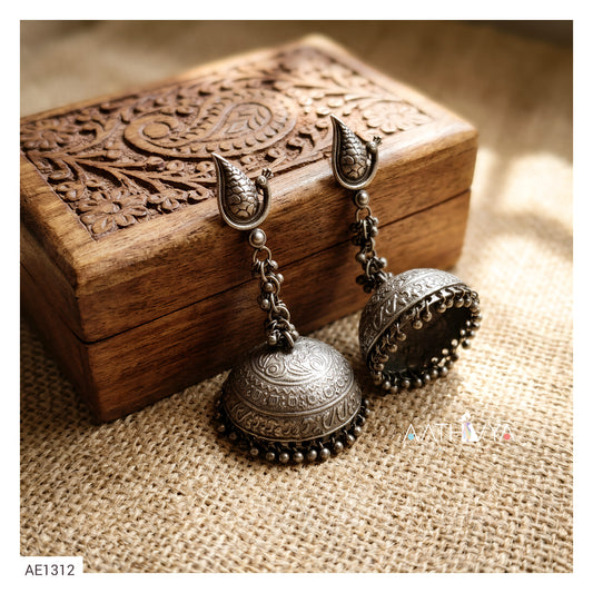 A pair of ornate silver-toned Indian jhumka earrings with detailed filigree work and bead borders, displayed on a carved wooden box over a burlap surface.