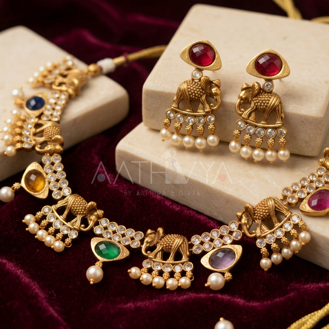 Gold-toned Indian jewelry set featuring a necklace and matching earrings with elephant motifs, pearl bead drops, and colorful gemstone accents, displayed on cream blocks over a dark red velvet background.